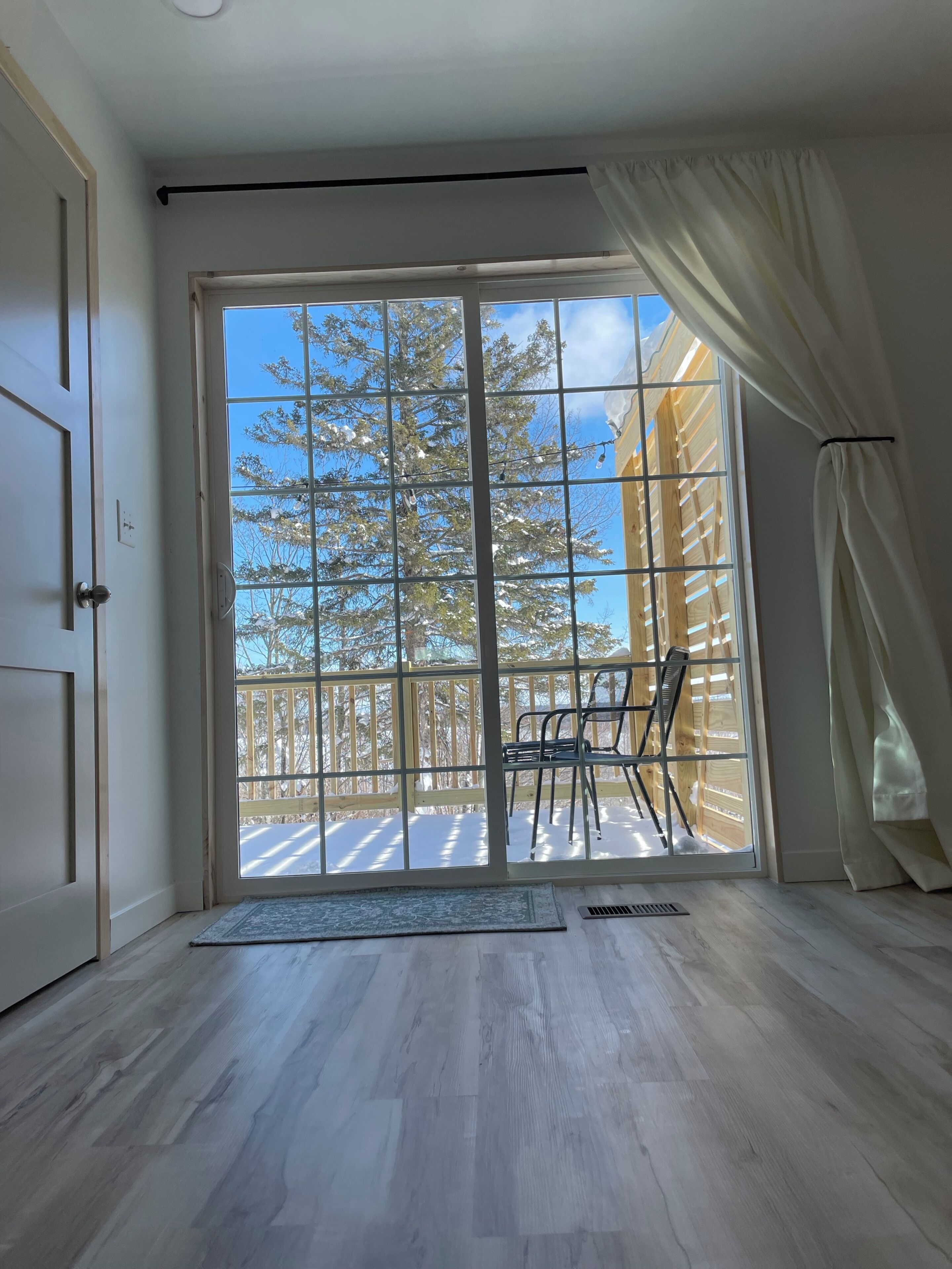 A room with a large sliding glass door leading to a wooden deck, a chair, and a rug on the floor.