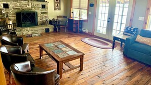 Un salon chaleureux avec une cheminée en pierre, des fauteuils en cuir, une table basse en bois et une vue sur l’extérieur par une porte.
