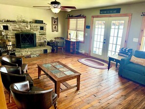 A cozy living room with a stone fireplace, leather chairs, a wooden coffee table, and a view of the outdoors through a door.