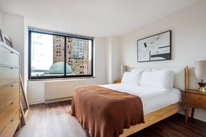 Ein Hotelzimmer mit einem Bett, einem Nachttisch, einer Lampe, einem Fenster mit Blick auf Gebäude und einem Wandbild.