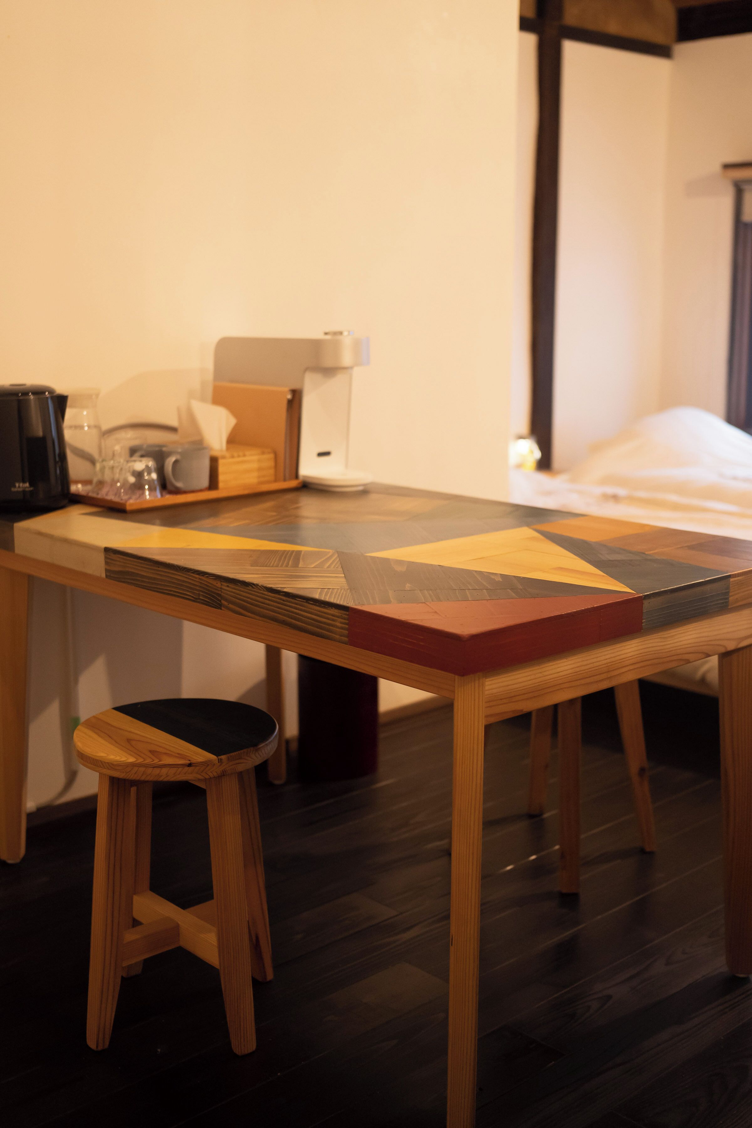 Una mesa de madera con tablero de diseño geométrico, una cafetera, un vaso y una caja de pañuelos.