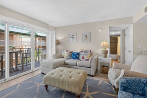 Ein Wohnzimmer mit einer Eckcouch, einem Hocker und einem kleinen Tisch mit Lampe. Es gibt einen Balkon mit Blick auf ein Gebäude mit begrünten Dächern.