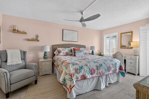 Ein Schlafzimmer mit einem Bett, einem Sessel, einem Nachttisch, einer Lampe, einem Deckenventilator und einem Fenster mit Fensterläden.