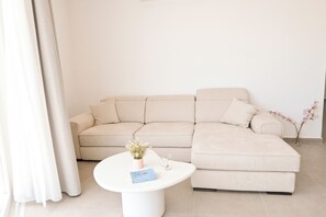 A modern living room with a beige sectional sofa, a white coffee table, and a vase with flowers. - Modern Seaside Apart in Marsalforn - 6B, Marsalforn Iz-Zebbug Gozo, Malta (Marsalforn Iz-Zebbug Gozo)