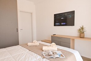 A hotel room with a bed, a bedside table with a remote control, a wall-mounted TV, and a small potted plant. - Modern Seaside Apart in Marsalforn - 6B, Marsalforn Iz-Zebbug Gozo, Malta (Marsalforn Iz-Zebbug Gozo)