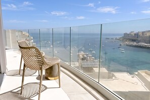 A balcony with a glass railing, a chair, and a view of the sea with boats and buildings. - Modern Seaside Apart in Marsalforn - 6B, Marsalforn Iz-Zebbug Gozo, Malta (Marsalforn Iz-Zebbug Gozo)