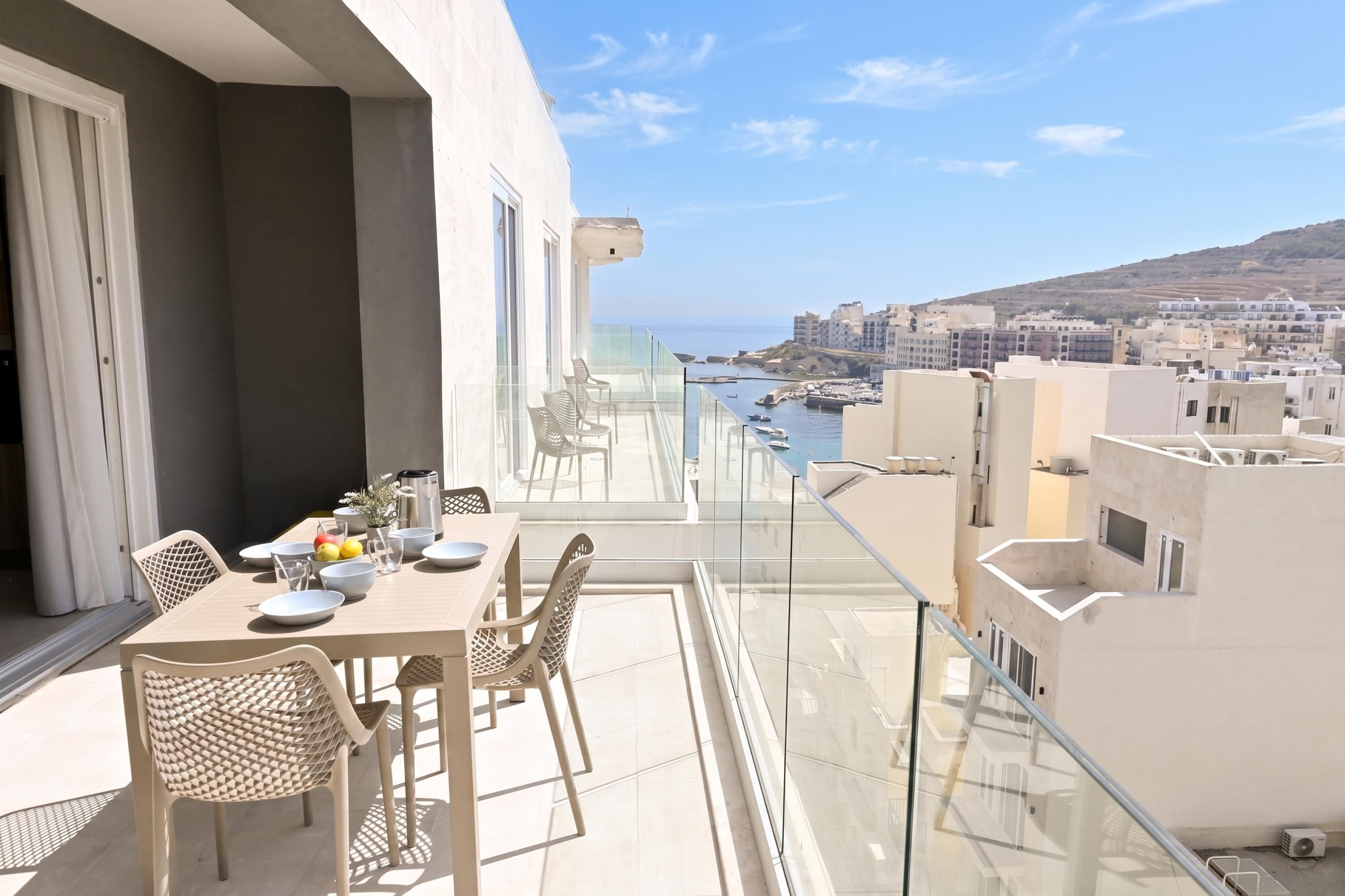 Varanda com mesa posta para refeições, vista para um horizonte costeiro com edifícios e barcos.