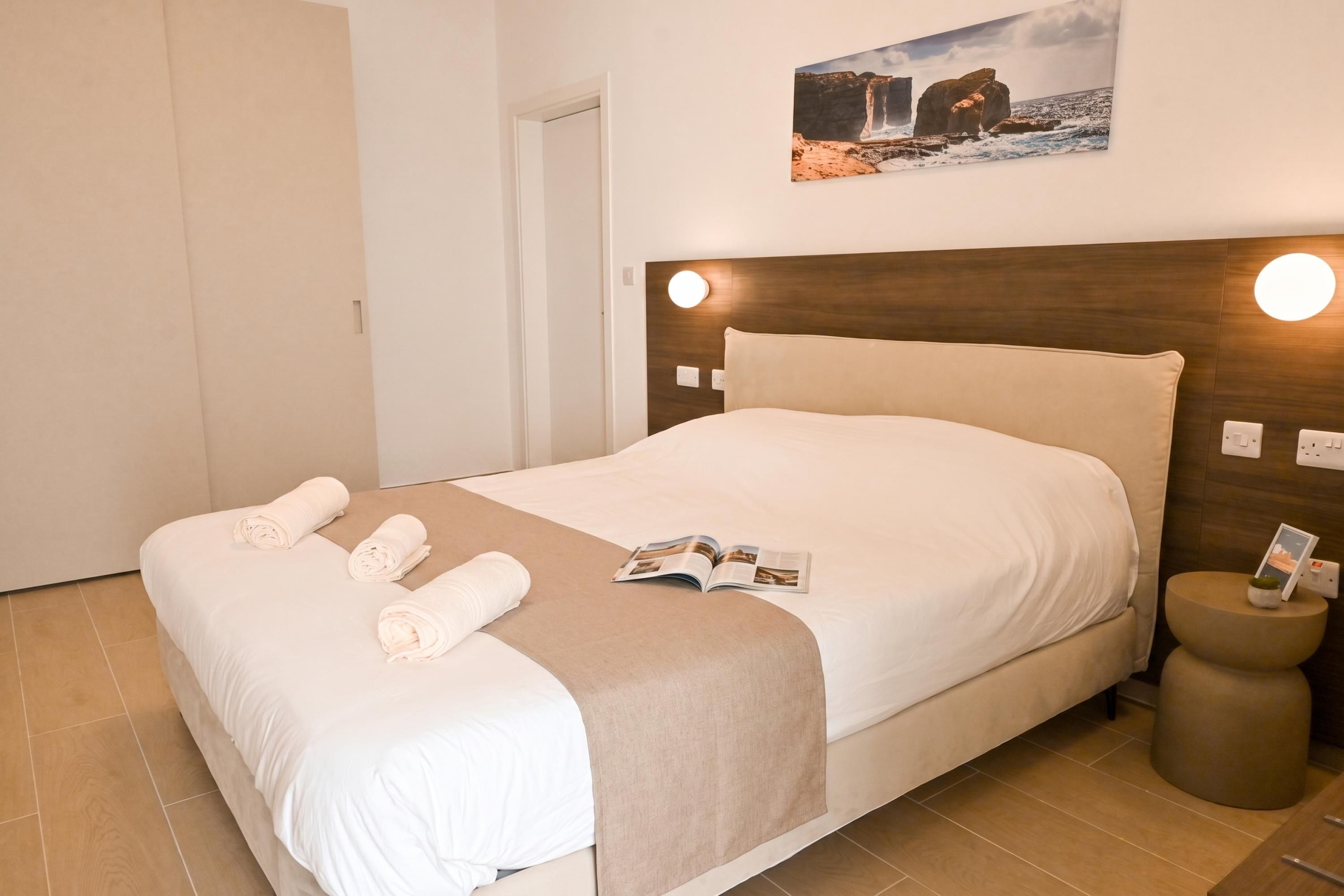 Una cama bien hecha con sábanas blancas y un plaid color beige, una revista sobre la cama, y una mesita redonda con una planta al lado.