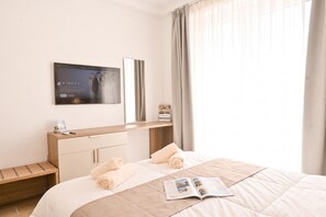 A hotel room with a bed, a bedside table, a TV mounted on the wall, and a window with curtains. - Modern Seaside Apart in Marsalforn - 6D, Marsalforn Iz-Zebbug Gozo, Malta (Marsalforn Iz-Zebbug Gozo)