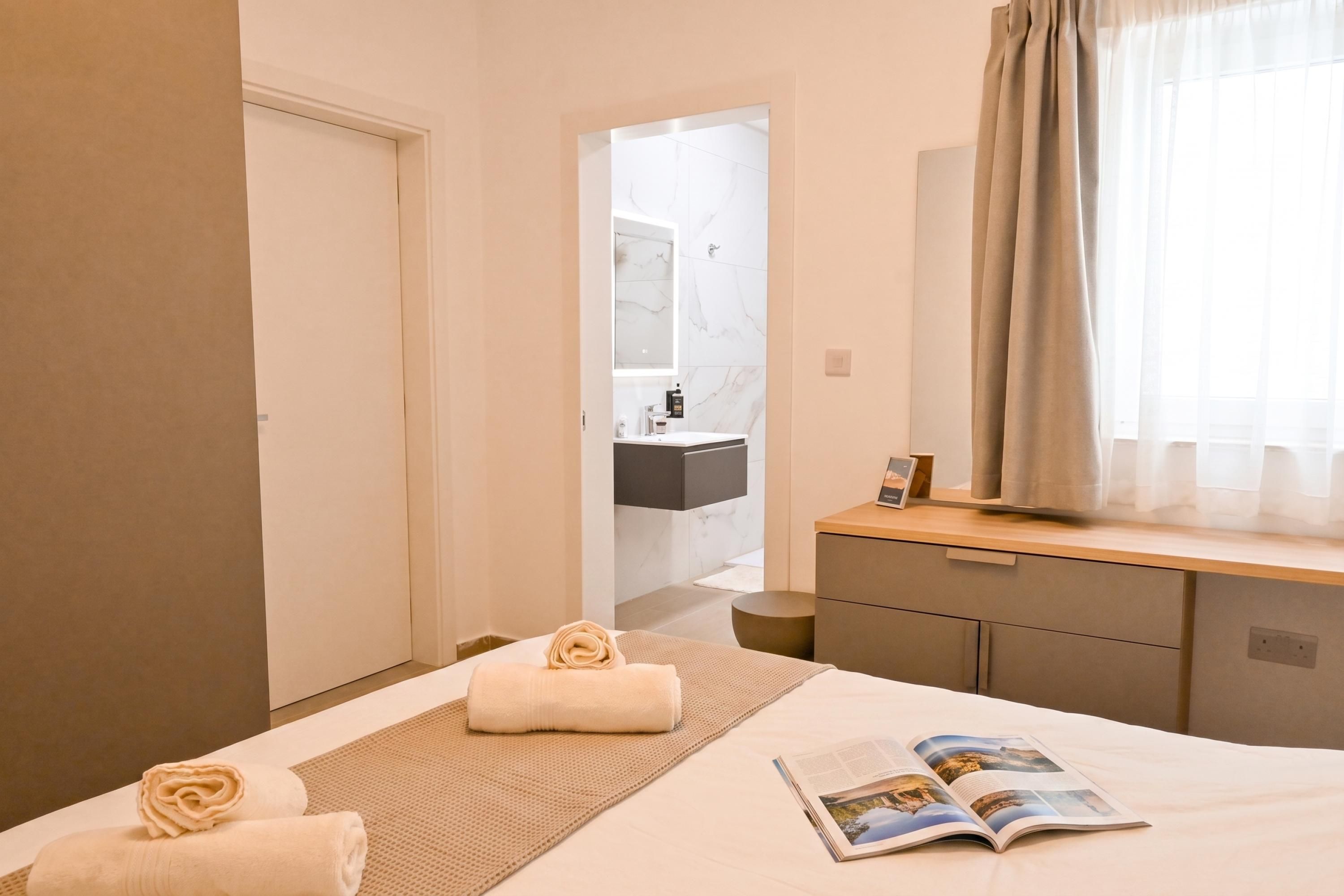Une chambre d’hôtel avec un lit, une table de chevet, un miroir, une fenêtre avec des rideaux et une salle de bain visible à travers une porte ouverte.