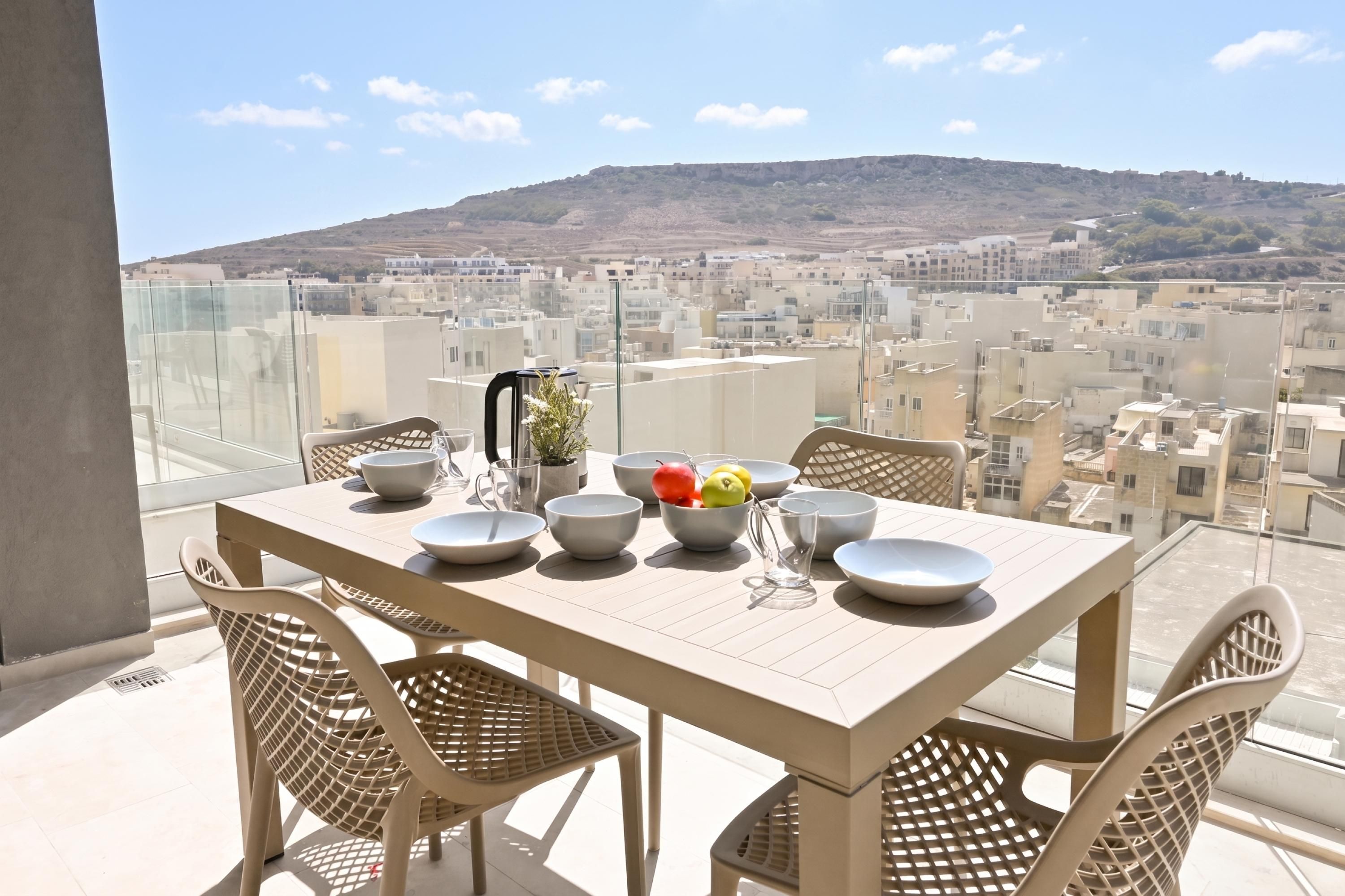 A balcony with a table set for dining, overlooking a cityscape with buildings and a hill.