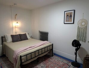 Ein Schlafzimmer mit einem Bett, einem Heizkörper, einer Wandleuchte, einem gerahmten Bild, einer Wanddekoration und einem Standventilator.