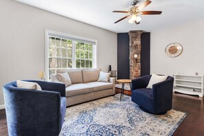 A living room with a sofa, two armchairs, a patterned rug, a ceiling fan, a brick accent wall, and a window with a view of greenery. - Hadlock House - NEW! Home in Southwest Harbor (Southwest Harbor)