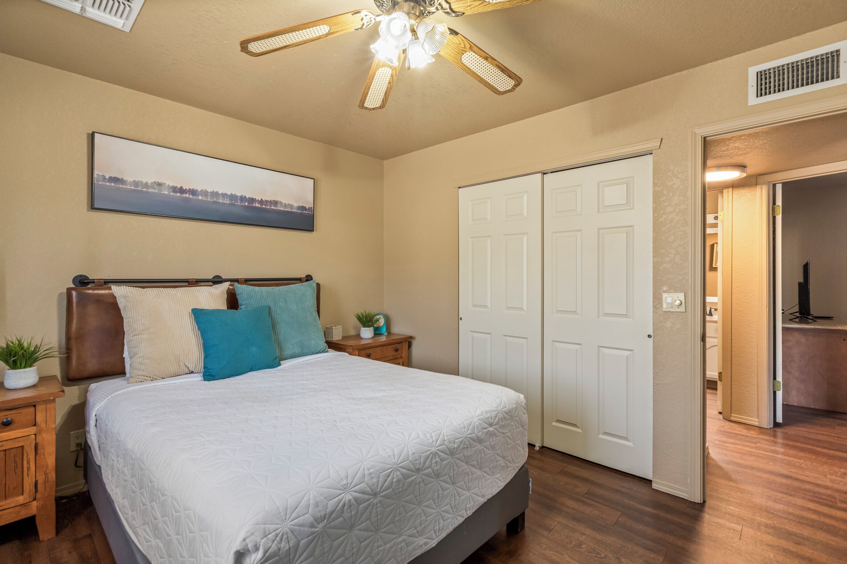 A bedroom with a bed, a ceiling fan, a nightstand, a bedside table, a wall-mounted clock, and a door.