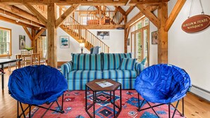 Een ruime kamer met een hoge plafond, houten balken, een gestreepte bank, twee blauwe fauteuils, een tafeltje en een vloerkleed.