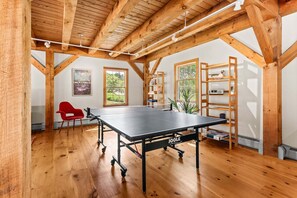 A ping pong table in a room with wooden beams, a red chair, and a bookshelf. - The Barn on the Bagaduce - NEW! Waterfront (Sedgwick)