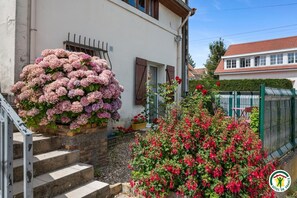 Ein Wohngebäude mit einer Treppe, die zu einer Tür führt, einer großen Blumenkomposition auf den Stufen und einem Garten mit roten Blüten.