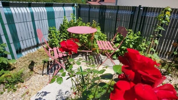 A garden area with a red table and chairs, surrounded by greenery and a fence. - Gîtes de France® - Between the Golf Course and the Sea (DIEPPE)
