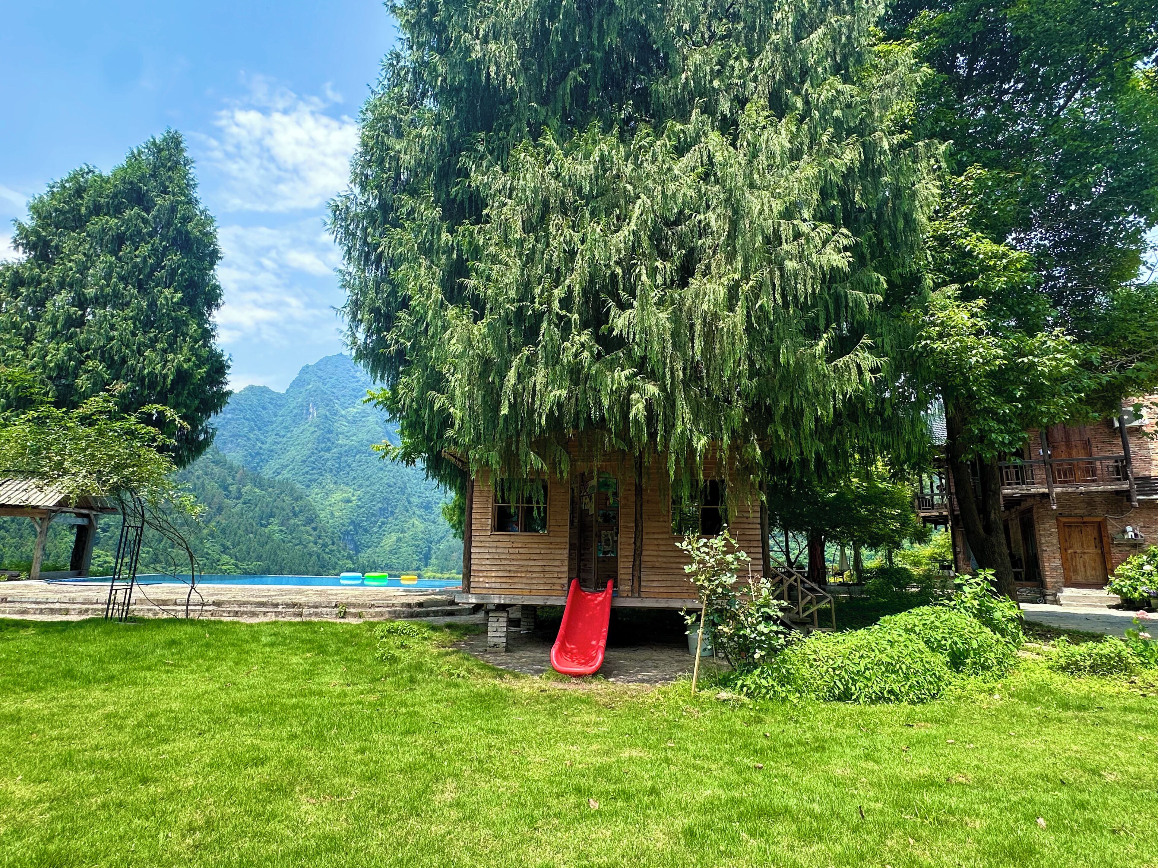 A rustic wooden cabin with a red slide, surrounded by lush greenery and a mountainous backdrop.