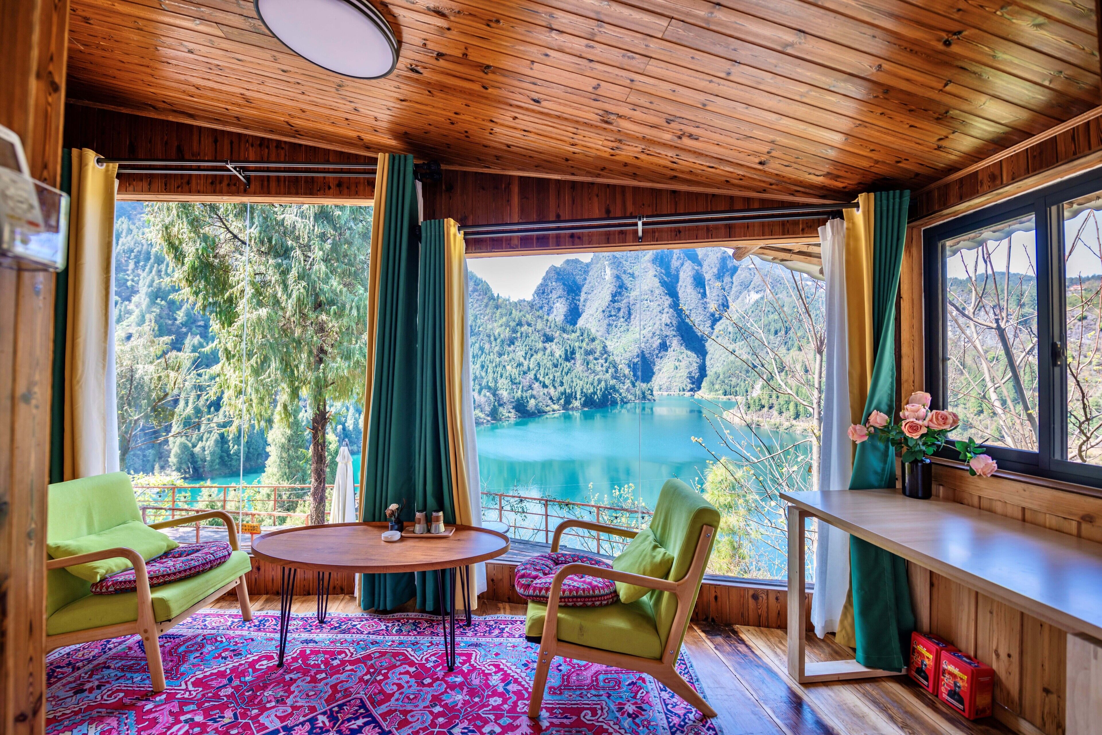 A room with a wooden ceiling, large windows with curtains, a round table, two chairs, and a view of a lake and mountains.