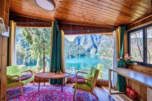 Una habitación con techo de madera, ventanas amplias con cortinas, una mesa redonda, dos sillas y vistas a un lago y montañas.