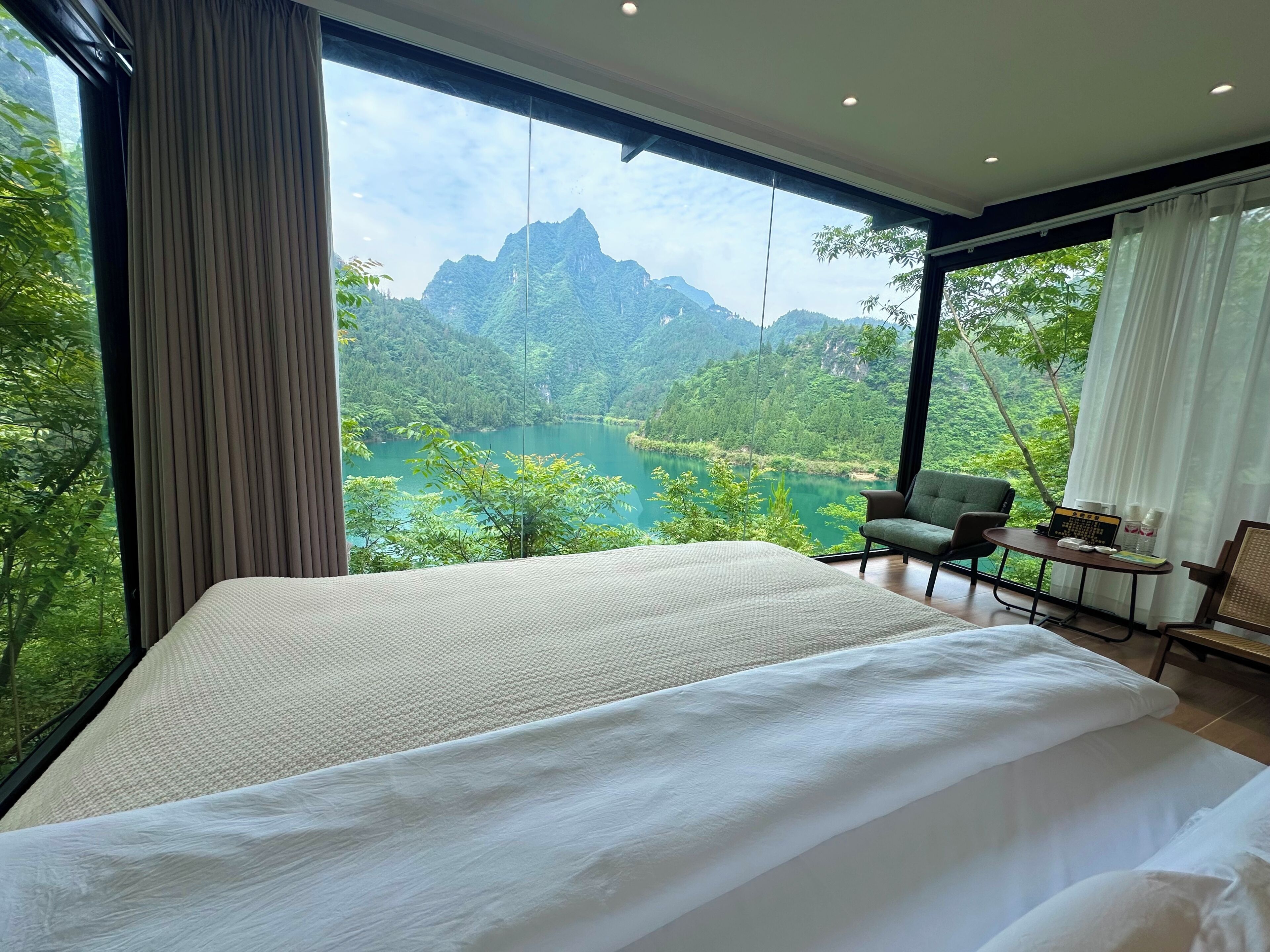 A bedroom with a large bed, a chair, a small table, and a view of a lake and mountains.