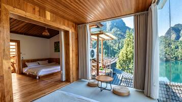 A wooden cabin with a bed, a small table, and a view of a lake and mountains.