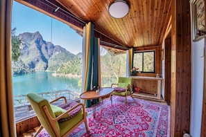 Una habitación con vistas a un lago y montañas, paredes de madera, una mesa redonda y una silla verde.