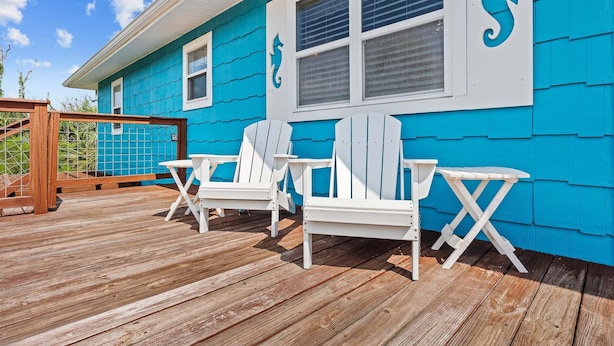 Eine Terrasse mit weißen Adirondack-Stühlen und einem Tisch, ein blaues Haus mit weißem Zierrahmen und ein Holzzaun.