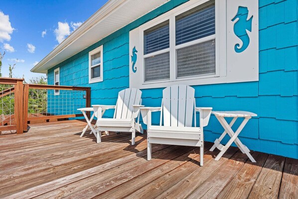 Una terraza con sillas Adirondack blancas y una mesa, una casa azul con detalles blancos y una valla de madera.