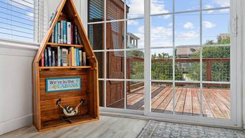 A corner with a wooden bookshelf, a hammock, and a view of a wooden deck through large windows.
