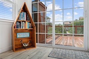 A corner with a wooden bookshelf, a hammock, and a view of a wooden deck through large windows.