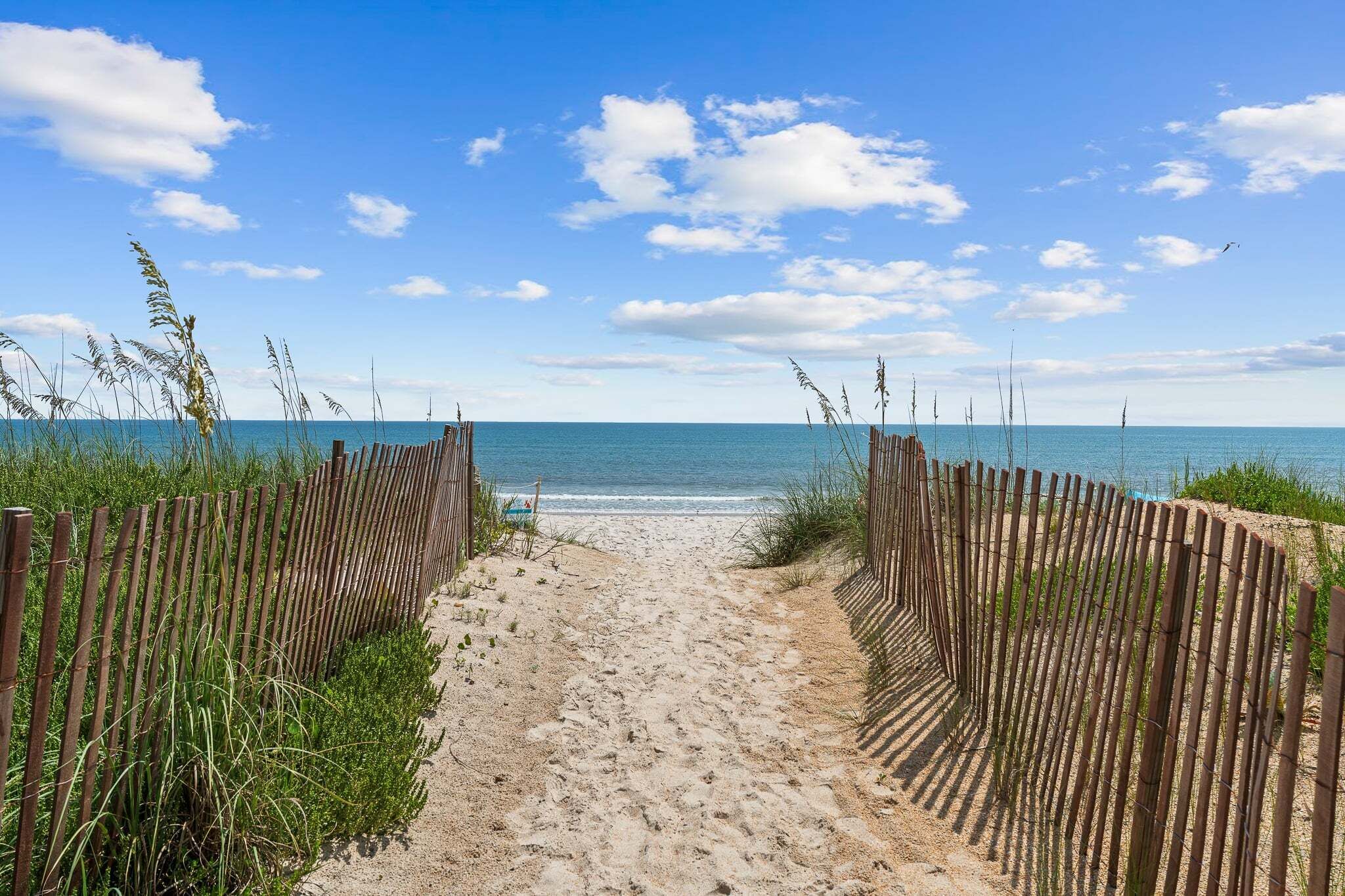 A sandy beach path leading to the sea, flanked by wooden fences and tall grasses.