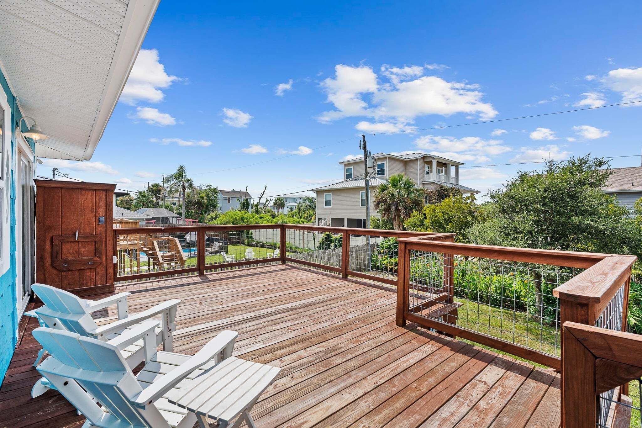 A wooden deck with a railing, white lounge chairs, and a view of a residential area with houses and greenery.