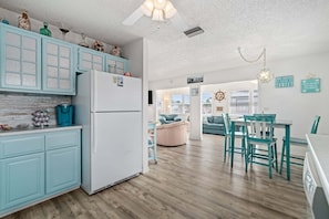 Una cocina con gabinetes azul claro, un refrigerador blanco y una zona de comedor con sillas color esmeralda y una mesa.