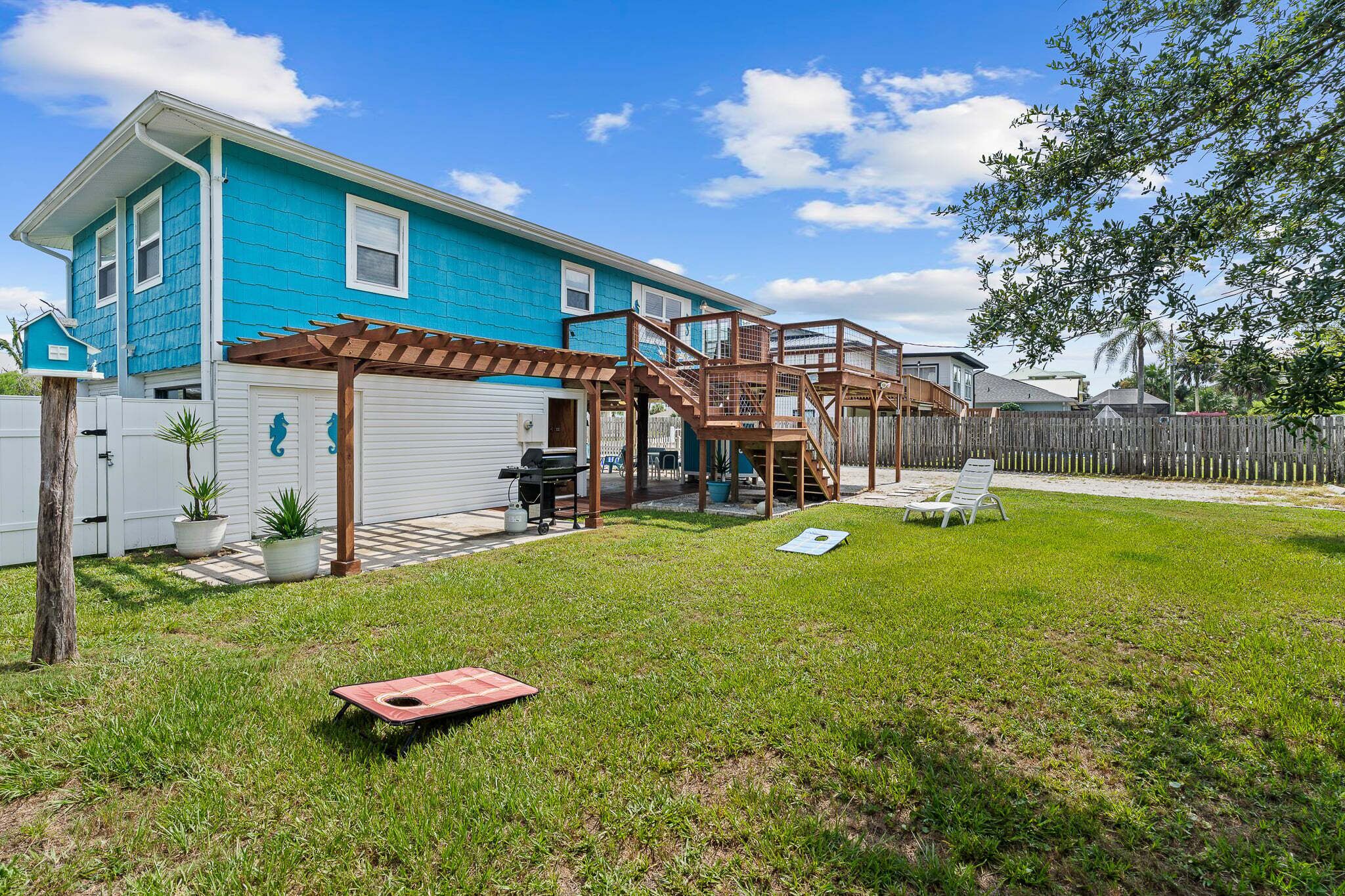 A residential backyard with a blue house, a wooden deck, and a covered patio area.
