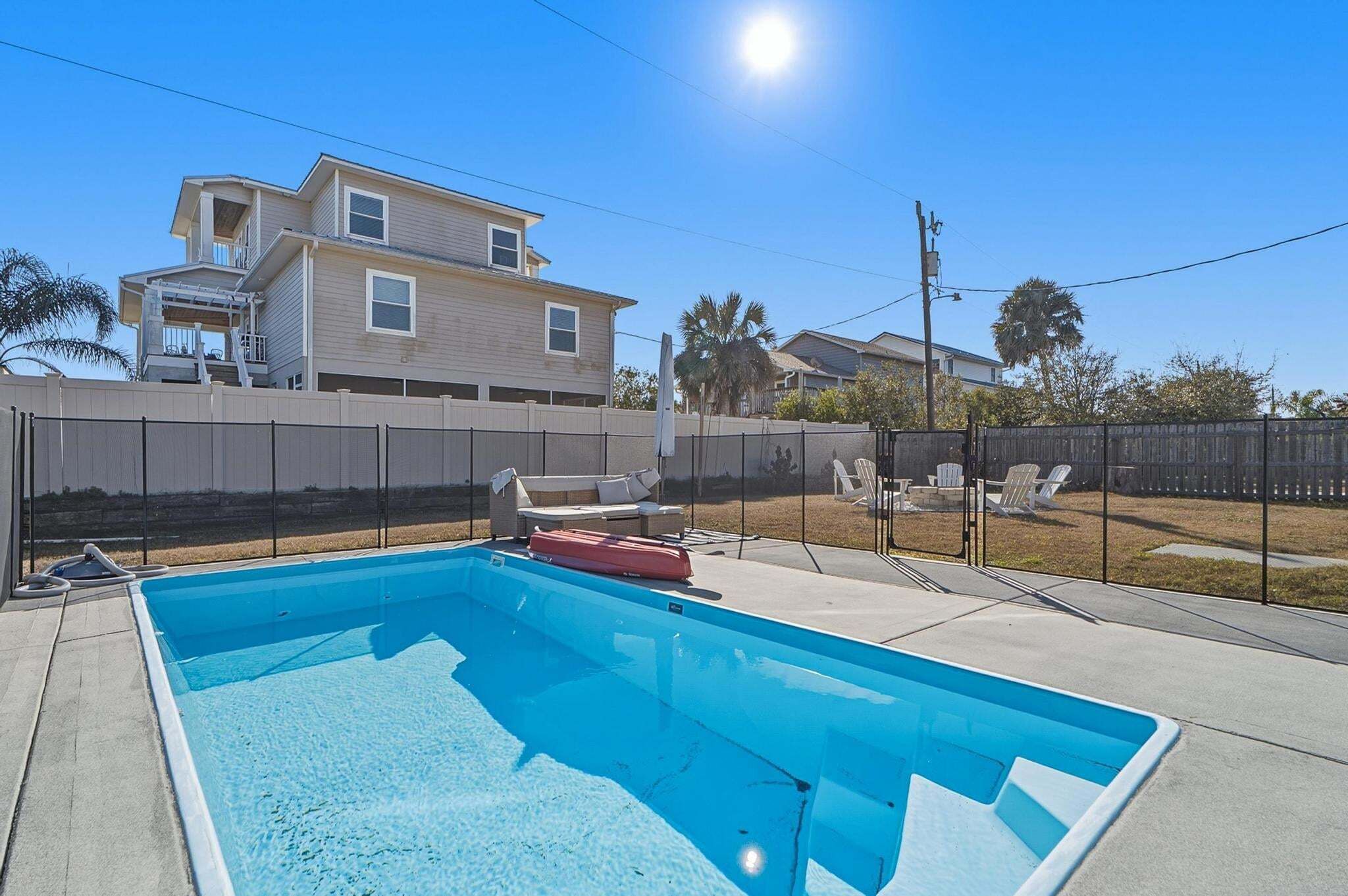 A rectangular swimming pool with a clear blue water surface, surrounded by a concrete deck. There are lounge chairs and a small table with a red object on it.