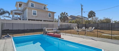 A rectangular swimming pool with a clear blue water surface, surrounded by a concrete deck. There are lounge chairs and a small table with a red object on it.