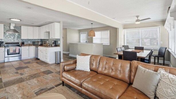 A spacious living area with a leather sofa, a dining table with chairs, a kitchen with white cabinets, and a ceiling fan.
