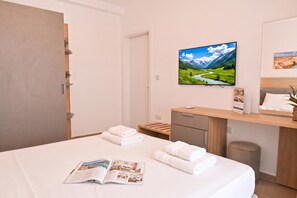 A hotel room with a bed, a desk, a chair, a small trash bin, a wall-mounted TV, and a magazine on the bed. - Modern Seaside Apart in Marsalforn - 5C, Marsalforn Iz-Zebbug Gozo, Malta (Marsalforn Iz-Zebbug Gozo)