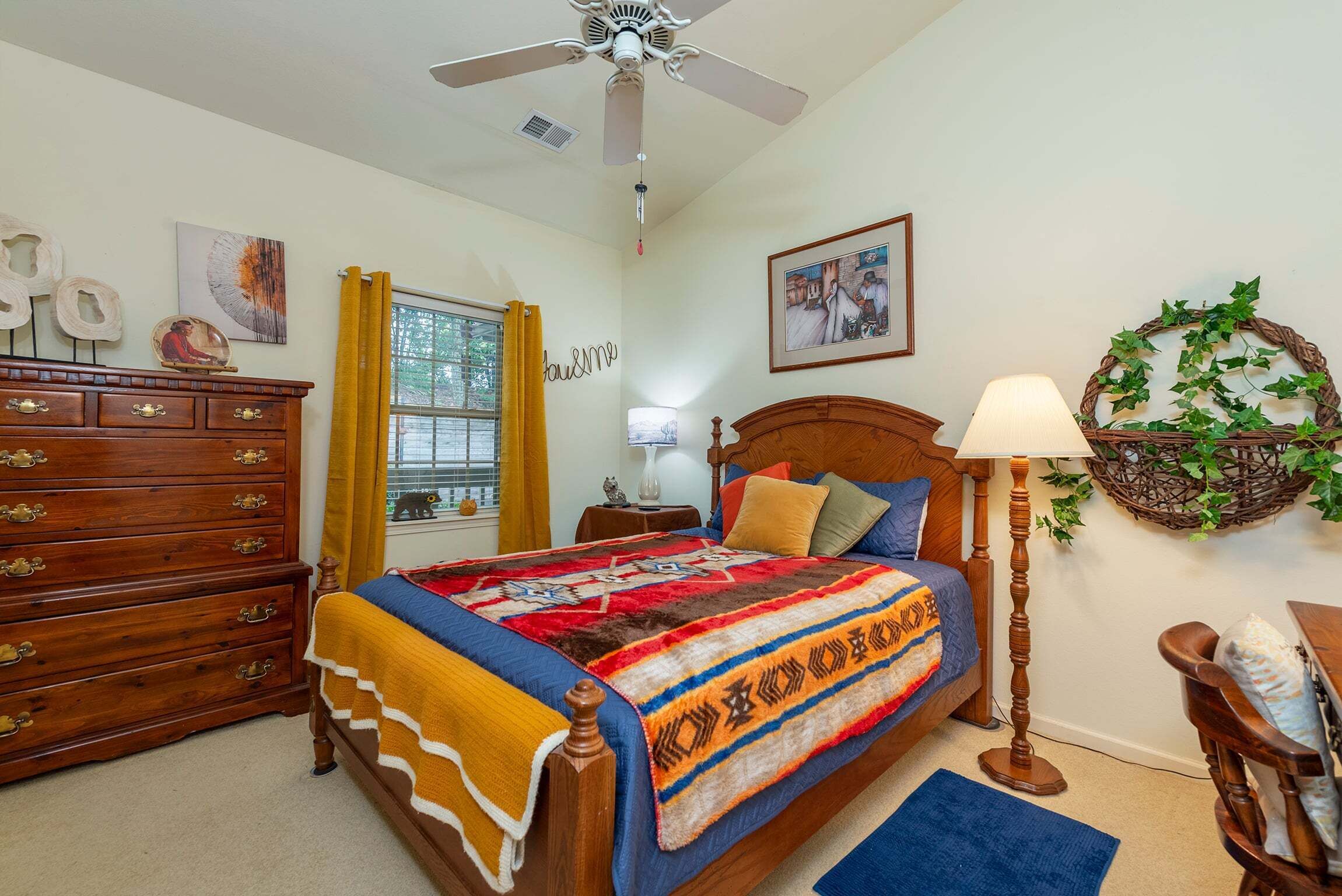 A bedroom with a bed, dresser, ceiling fan, window with curtains, wall art, and a lamp.