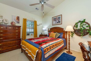 Un dormitorio con cama, cómoda, ventilador de techo, ventana con cortinas, decoración en la pared y una lámpara.
