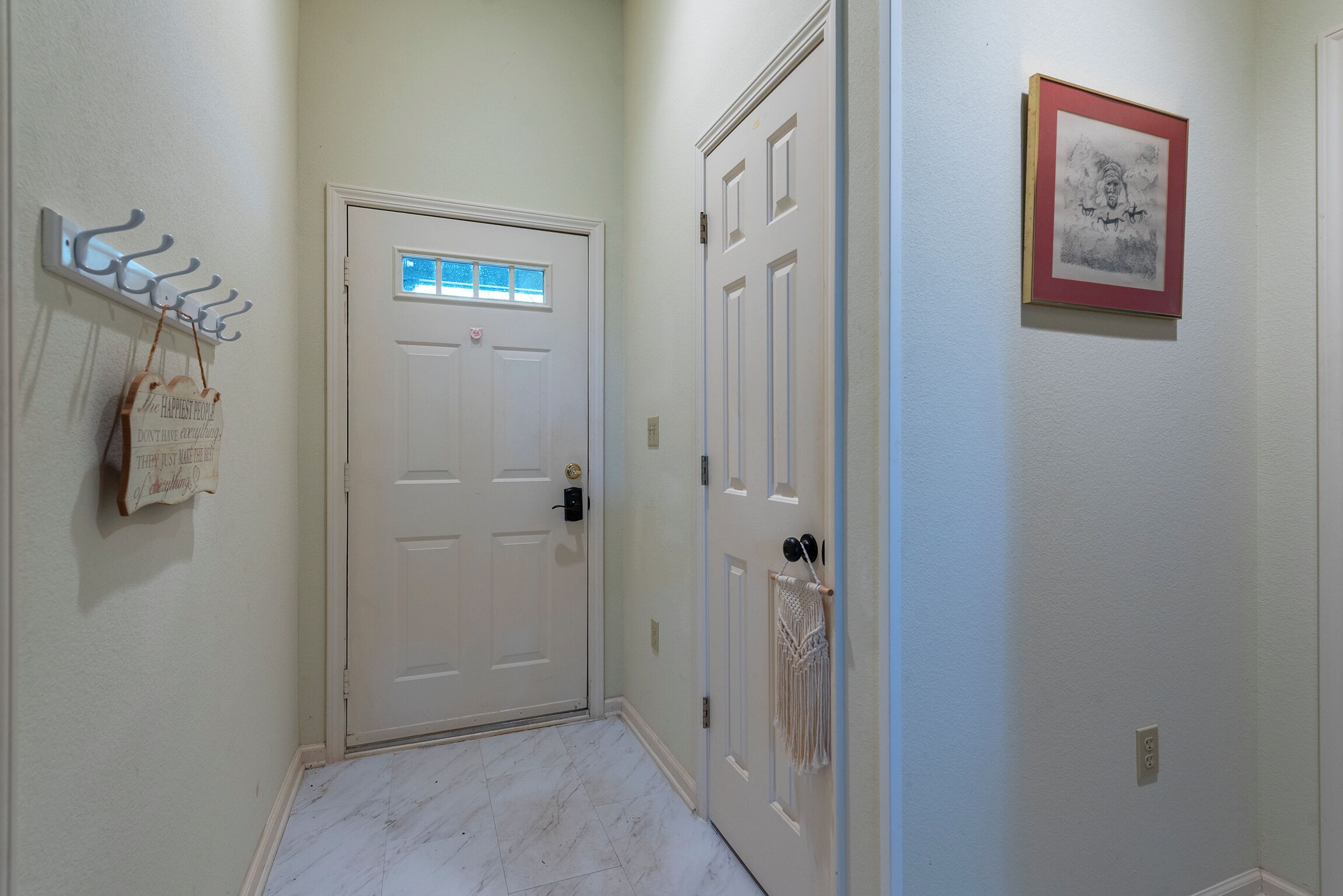 A hallway with a white door, a coat rack with a sign, a macrame wall hanging, and a framed picture on the wall.