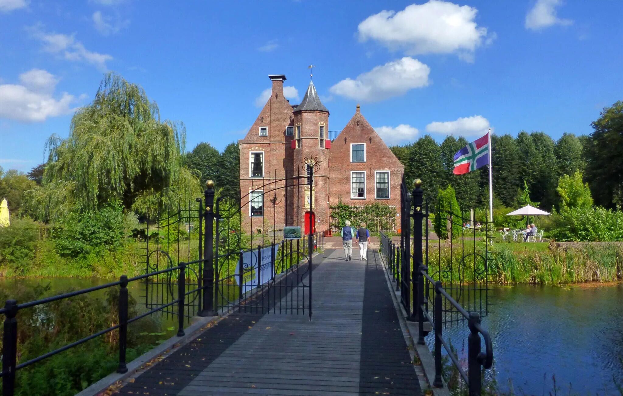 Un pont en bois mène à un bâtiment historique en briques, orné d’un drapeau, et est entouré de verdure et bordé par une étendue d’eau paisible.