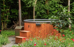 A hot tub with a wooden exterior and steps leading up to it, surrounded by a garden with various flowers.