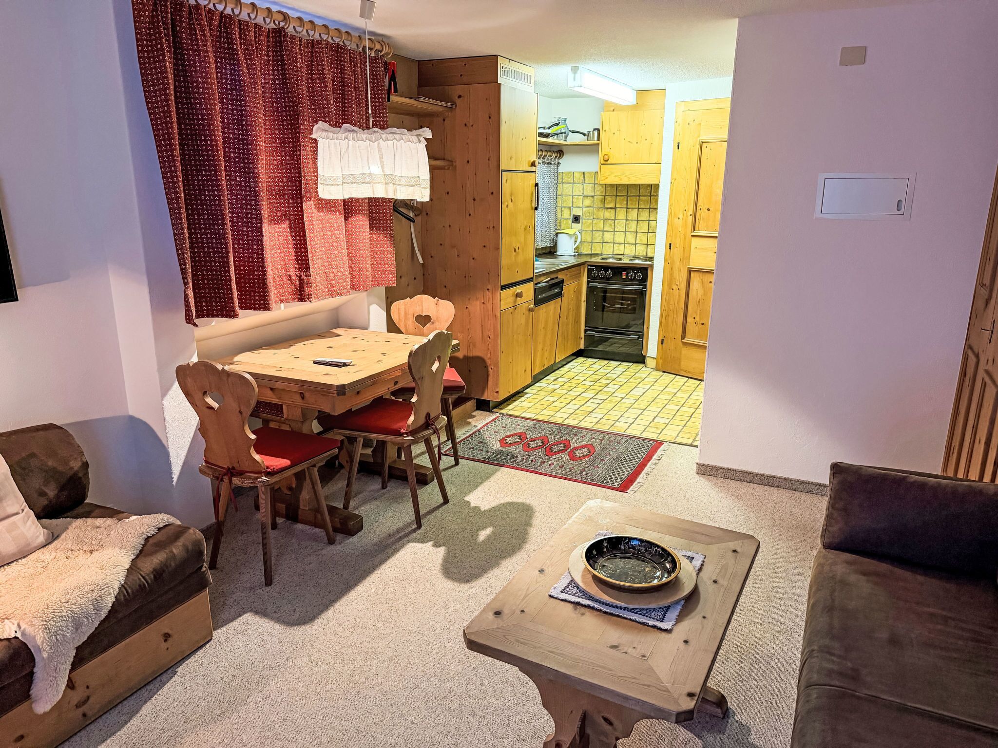 A cozy living room with a wooden dining table, chairs, and a sofa. A kitchen is visible through an open doorway.