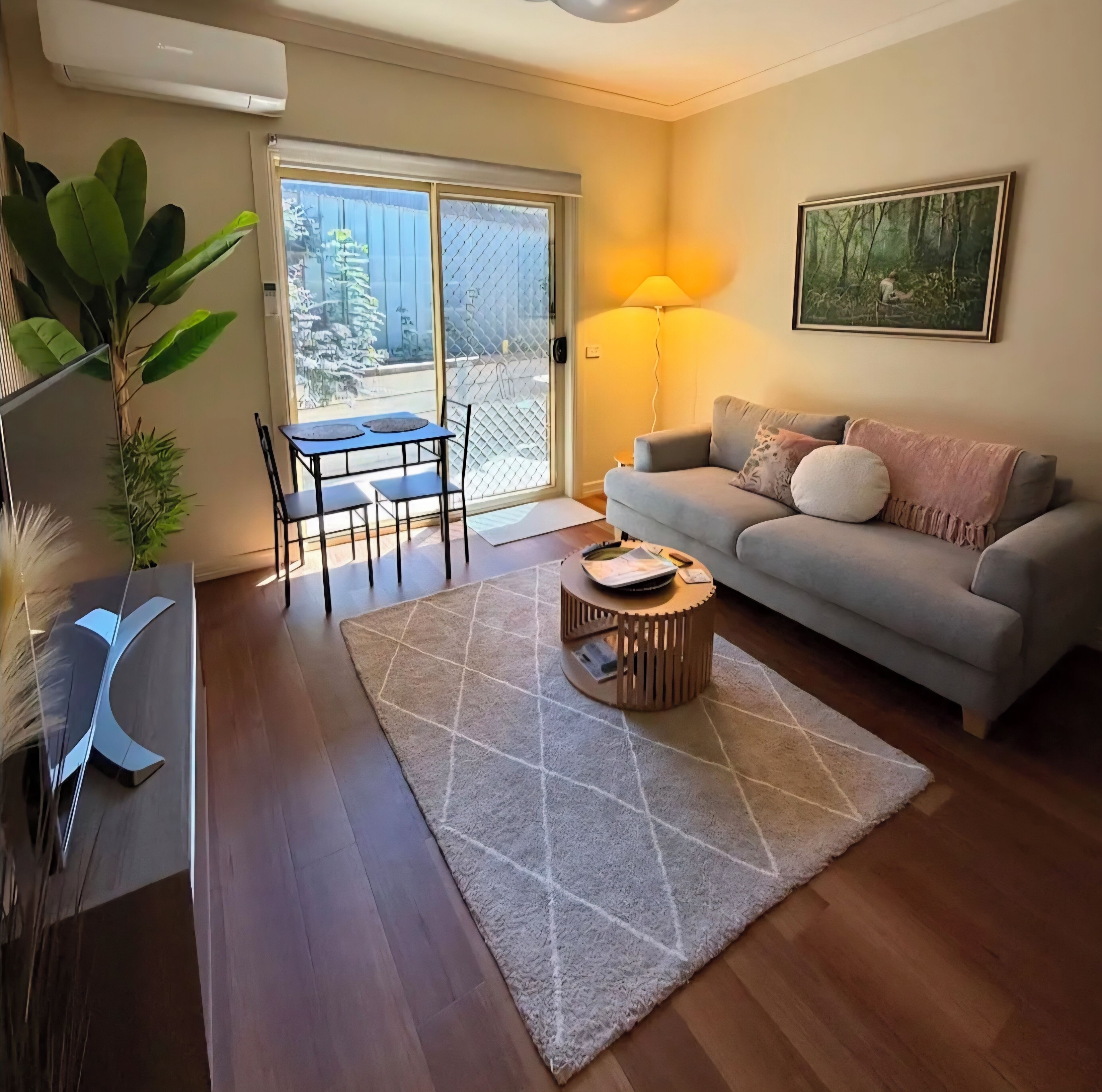 A living room with a sofa, a coffee table, a dining table with chairs, a large window, and a painting on the wall.