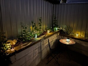 Un rincón al aire libre acogedor con una mesa y sillas, rodeado de plantas e iluminado con luz tenue.