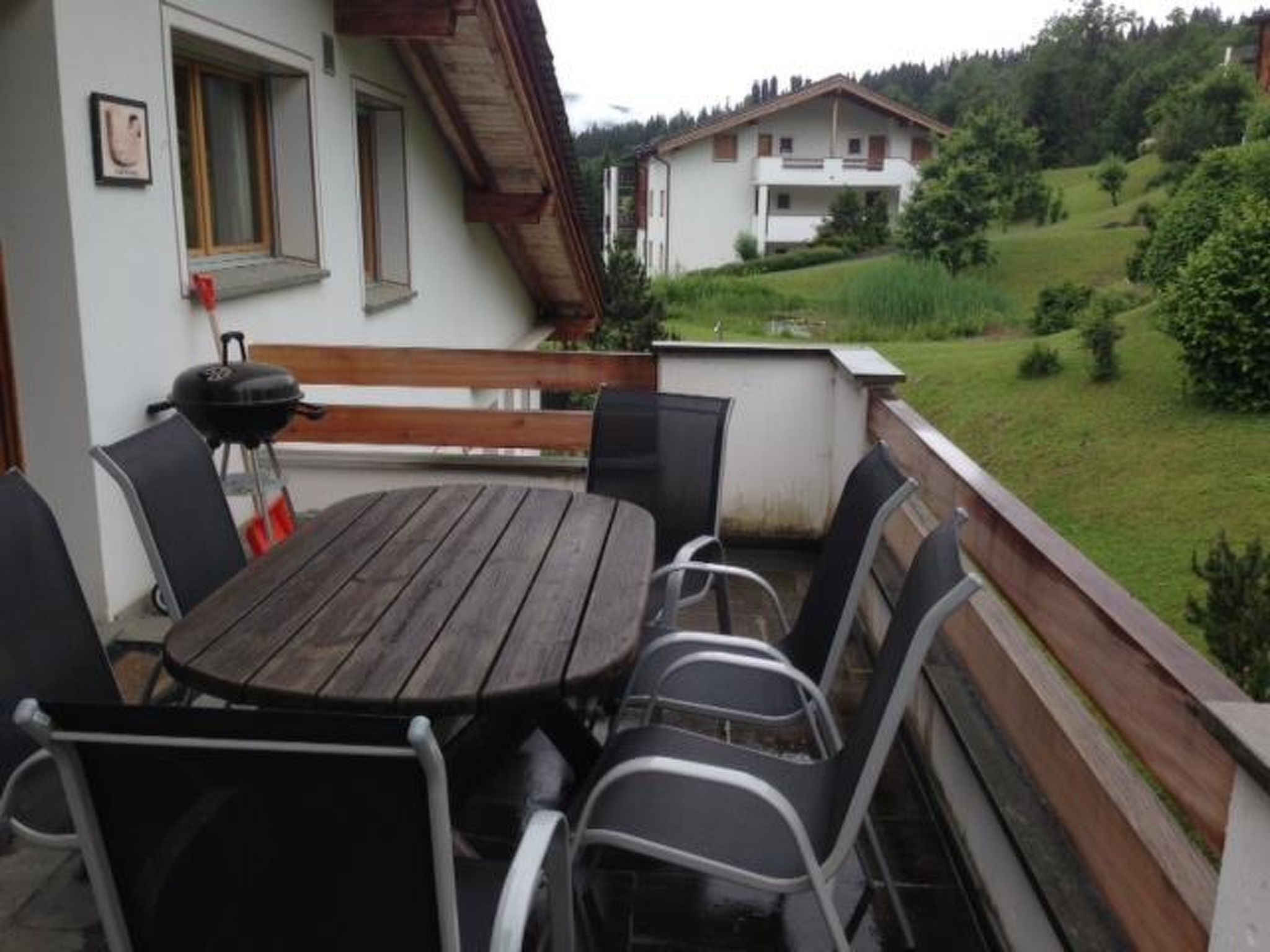 A patio with a round table and chairs, a barbecue grill, and a view of a house and greenery.