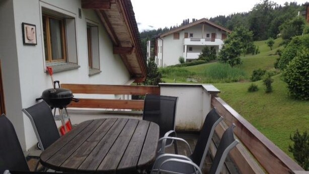 A patio with a round table and chairs, a barbecue grill, and a view of a house and greenery. - Mira Selva, (Flims Dorf), by Interhome (Flims)